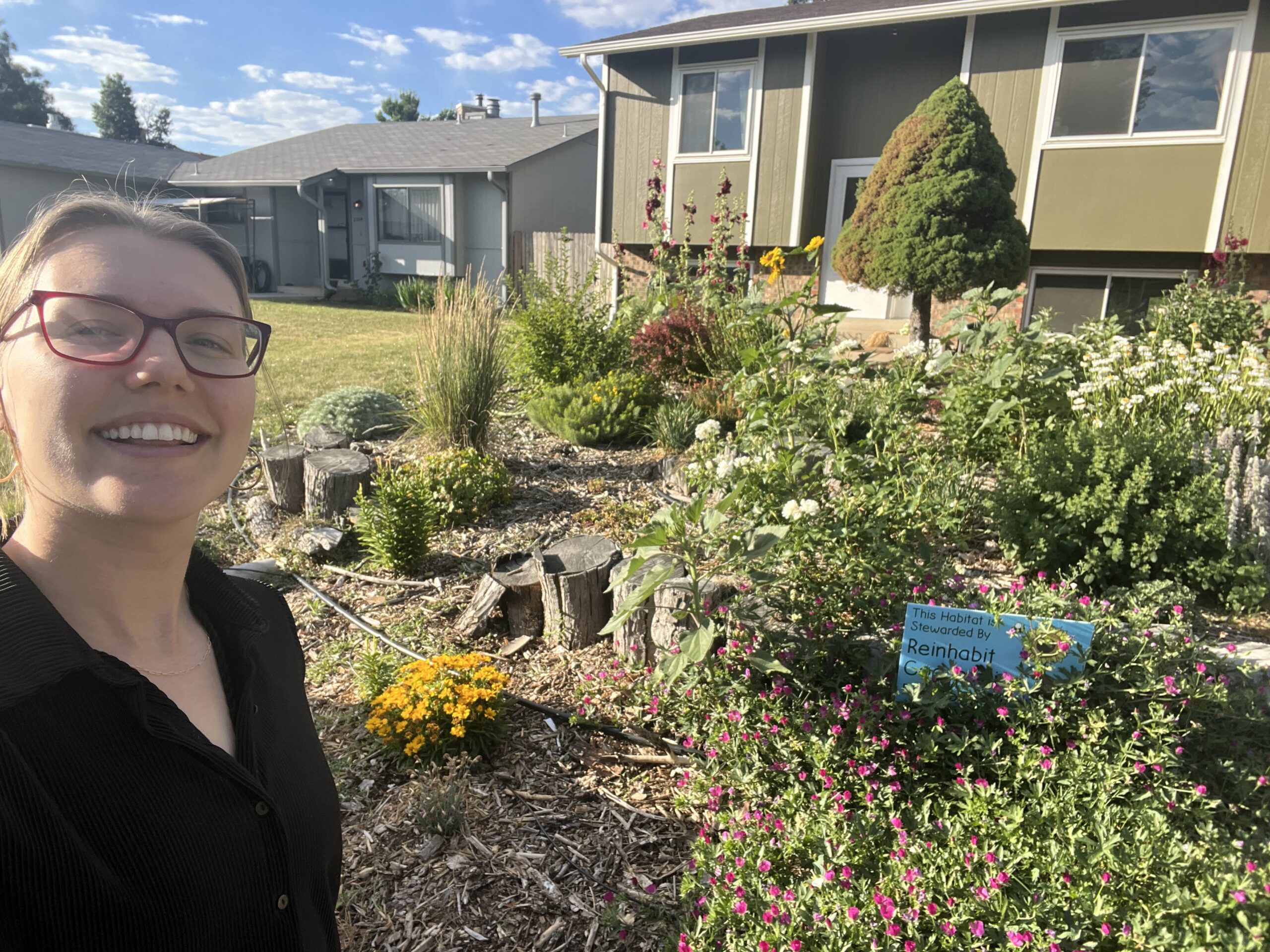 Woman with a black shirt and red glasses smiles next to her garden bed full of life and flowers.